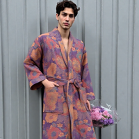 Man wearing a patterned robe holding flowers against a gray wall. (Purple)