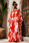A woman wearing a vibrant red, pink, and white floral print kaftan dress stands outdoors next to a rustic wooden door and a potted plant.