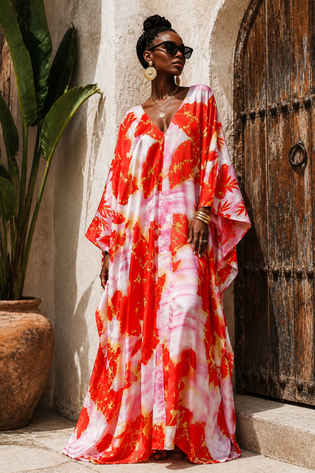 A woman wearing a vibrant red, pink, and white floral print kaftan dress stands outdoors next to a rustic wooden door and a potted plant.