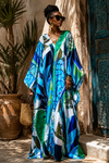 A woman wearing a vibrant blue, green, and white abstract print kaftan dress, standing outdoors in front of a rustic stone wall and wooden door.
