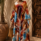 A woman wearing a long, flowing kaftan dress with a vibrant multicolor abstract print in blue, orange, brown, and cream, standing in front of a sunlit textured wall next to a large potted plant.