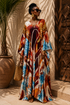 A woman wearing a long, flowing kaftan dress with a vibrant multicolor abstract print in blue, orange, brown, and cream, standing in front of a sunlit textured wall next to a large potted plant.