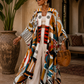 A woman stands in a warm-toned architectural setting wearing a long, open-front kaftan-style cardigan with bold abstract geometric patterns in orange, teal, black, white, and brown. She is also wearing white wide-leg pants, sunglasses, and carrying a woven straw bag.