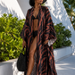 Woman in a black and brown patterned cover-up and black hat standing outdoors with greenery and a white architectural structure in the background.