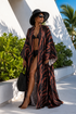 Woman in a black and brown patterned cover-up and black hat standing outdoors with greenery and a white architectural structure in the background.
