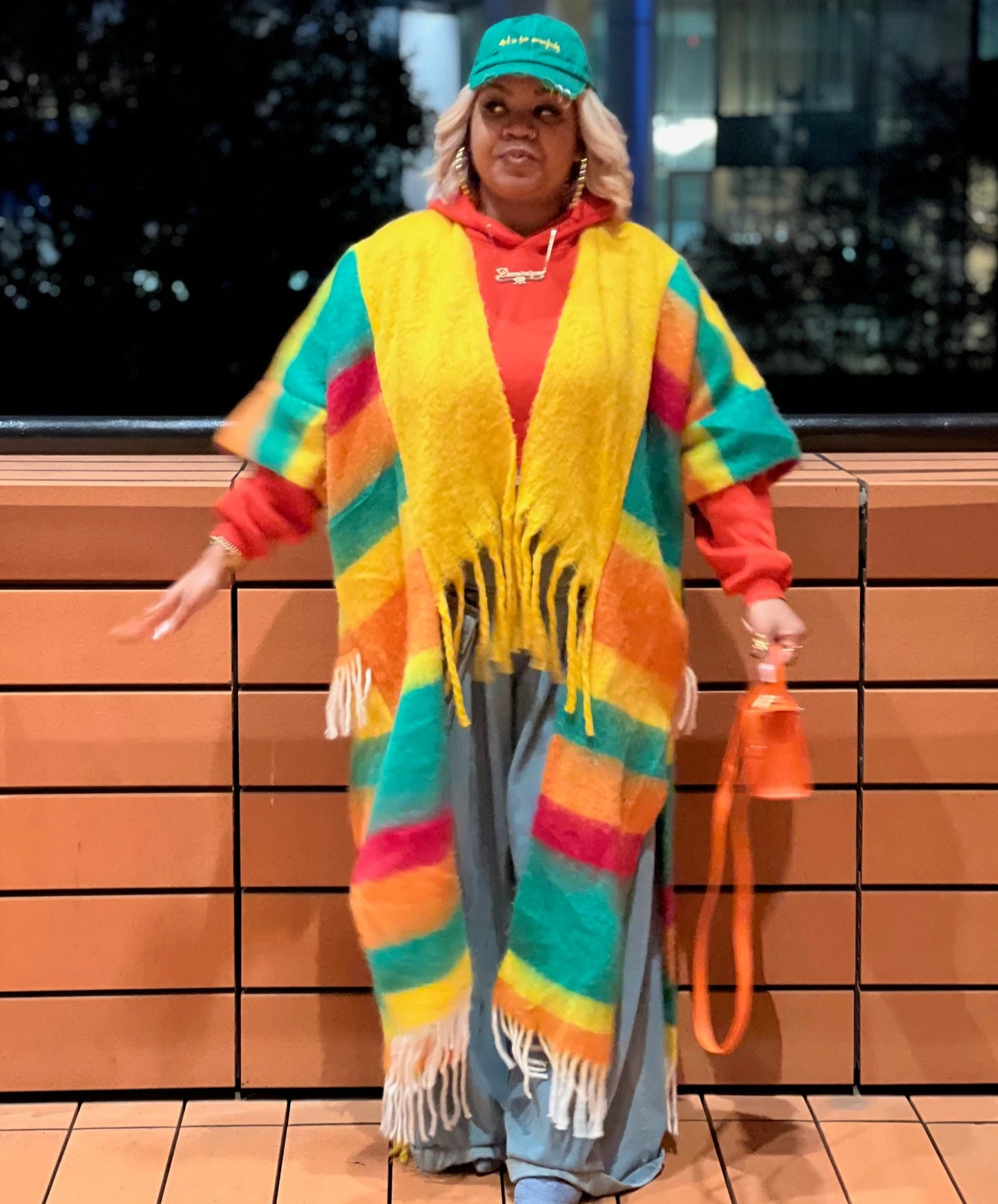 Person wearing a colorful striped cardigan and holding an orange bag against a brick wall. (Yellow)