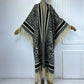 A long open-front poncho-style cardigan with wide sleeves, shown on a mannequin. The garment features a leopard print pattern in black, gray, and beige, with thick striped bands and long cream fringe along the edges and hem. (Black)