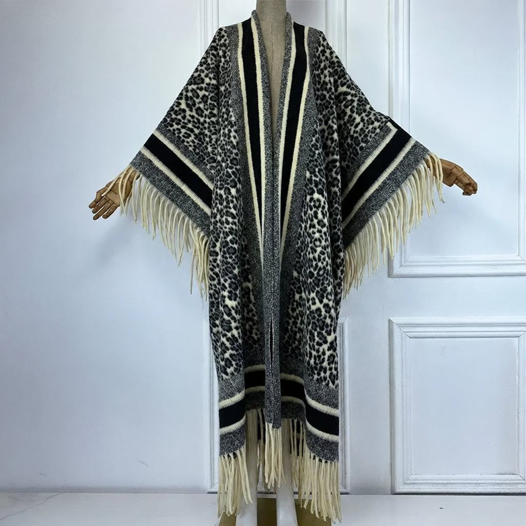 A long open-front poncho-style cardigan with wide sleeves, shown on a mannequin. The garment features a leopard print pattern in black, gray, and beige, with thick striped bands and long cream fringe along the edges and hem. (Black)
