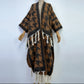 A brown and black patterned open-front cardigan or poncho with white braided fringe, displayed on a mannequin against a plain light background. (Brown)