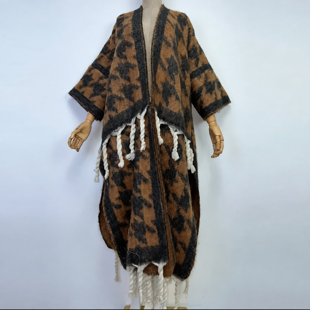 A brown and black patterned open-front cardigan or poncho with white braided fringe, displayed on a mannequin against a plain light background. (Brown)