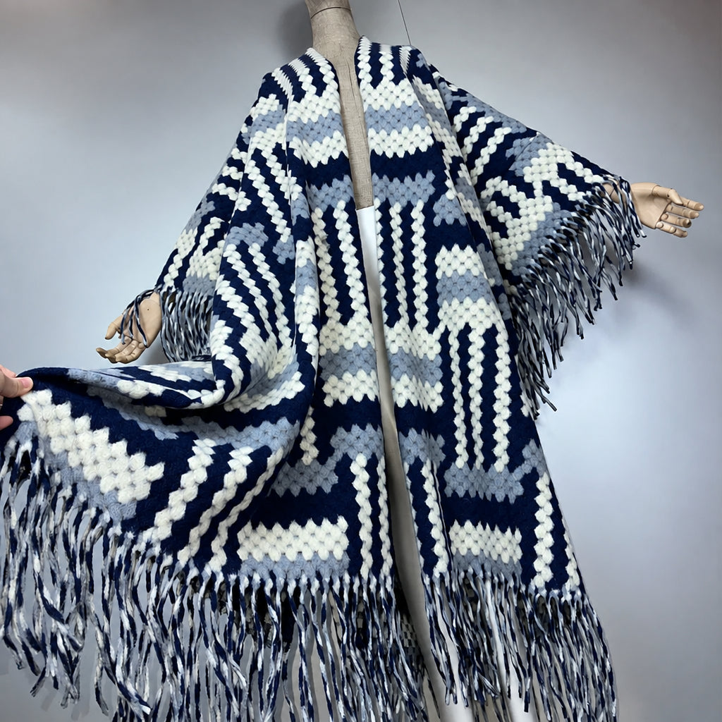 A chunky knit poncho sweater with a geometric pattern in navy blue, light blue, white, and gray, featuring long fringe on the hem and sleeves, displayed on a mannequin. (Grey)