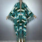 A crocheted open-front poncho or cardigan with wide sleeves, shown on a mannequin against a gray studio background. The garment has a bold geometric zigzag pattern in dark green, turquoise blue, and off-white, with long fringe trim along the hem and sleeves. (Green)