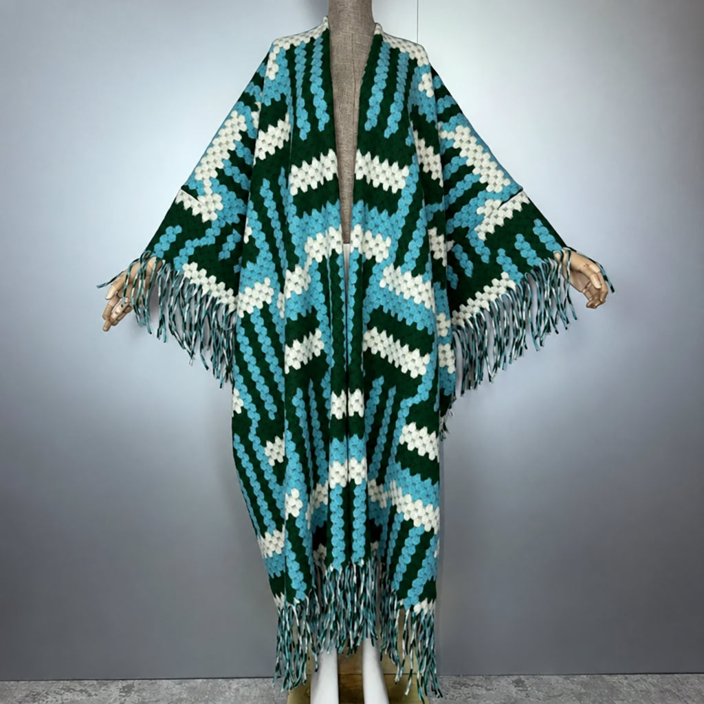 A crocheted open-front poncho or cardigan with wide sleeves, shown on a mannequin against a gray studio background. The garment has a bold geometric zigzag pattern in dark green, turquoise blue, and off-white, with long fringe trim along the hem and sleeves. (Green)