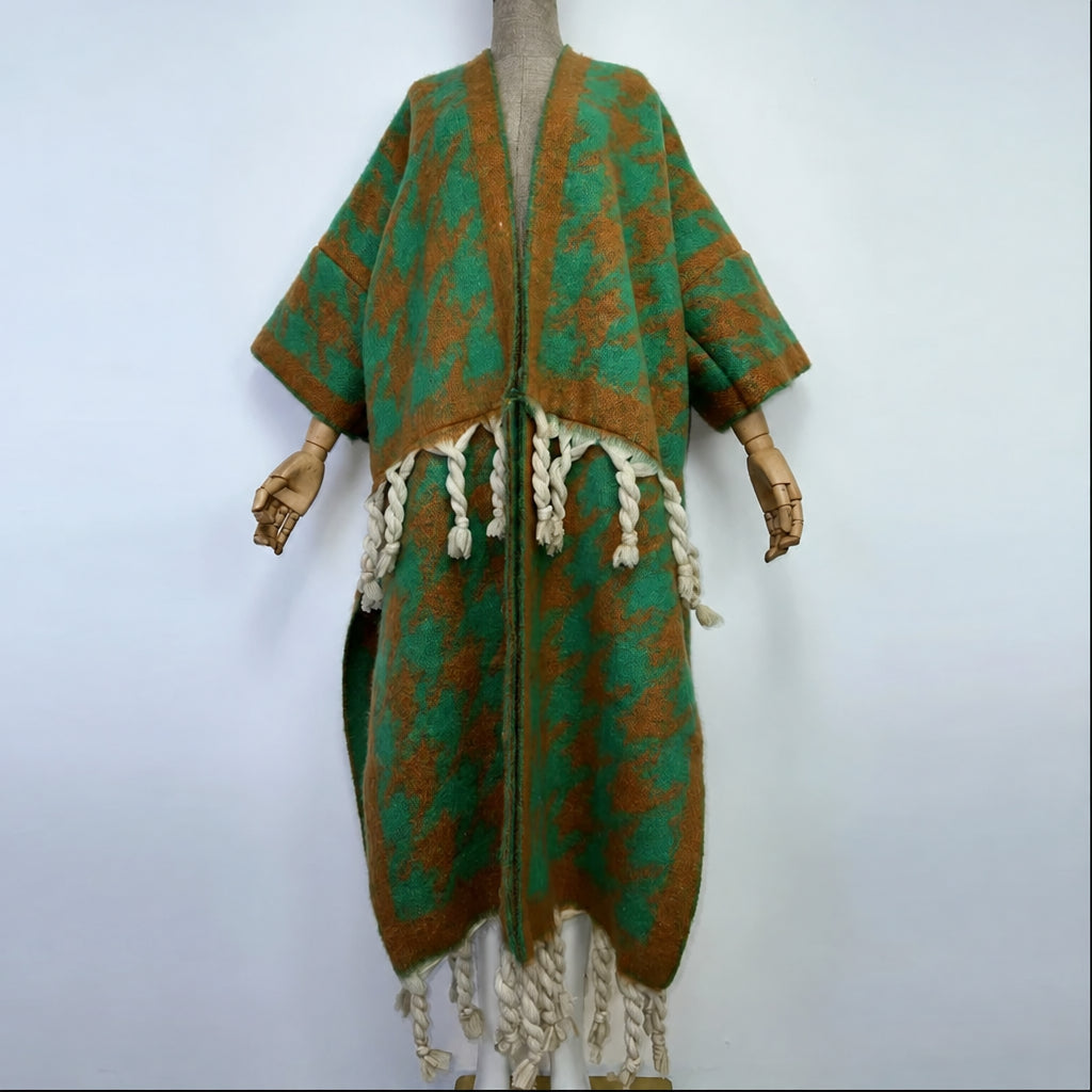 A green and brown patterned knit poncho with cream braided fringe, displayed on a mannequin with wooden hands against a plain light gray background. (Green)