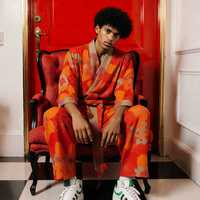 Person wearing an orange outfit with a patterned robe sitting on a red chair against a red wall. (Red)